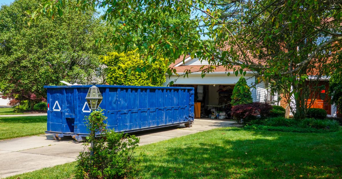 A large blue rental dumpster stationed on a residential driveway in front of a house with an open garage door.