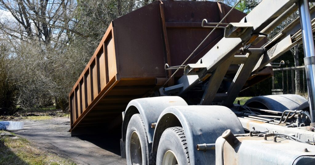 A delivery truck's hydraulic system drops off a large, brown roll-off dumpster on a dirt driveway in front of several trees.