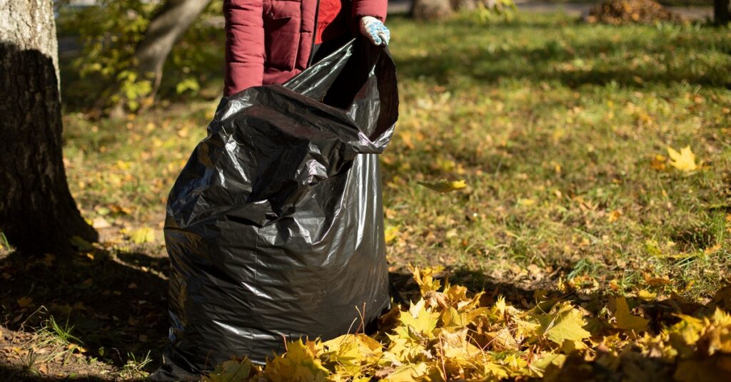 A woman wearing a red puffer jacket holds a large black trash bag open, standing near a pile of fallen leaves.