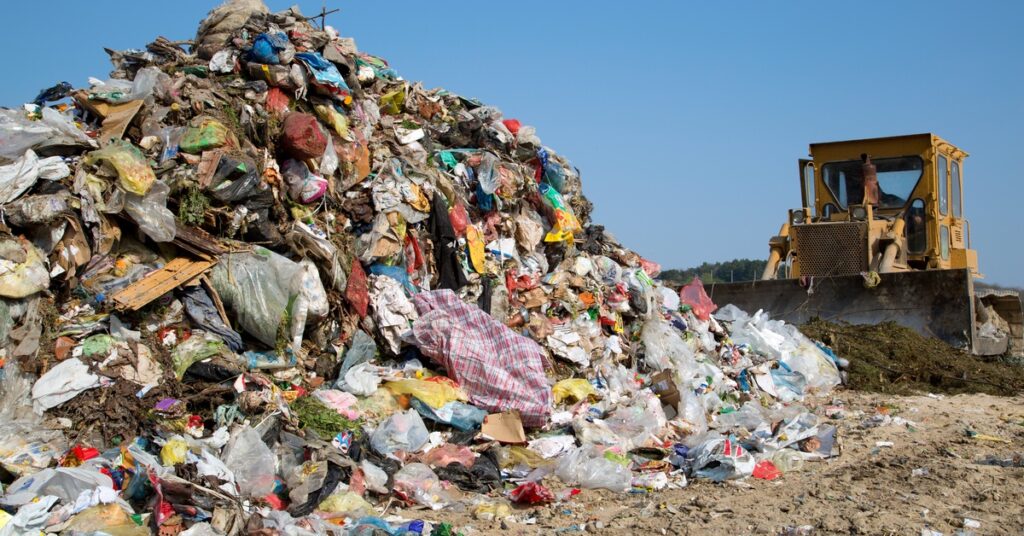 A yellow bulldozer pushes a pile of tree branches toward a large pile of assorted junk in a landfill.