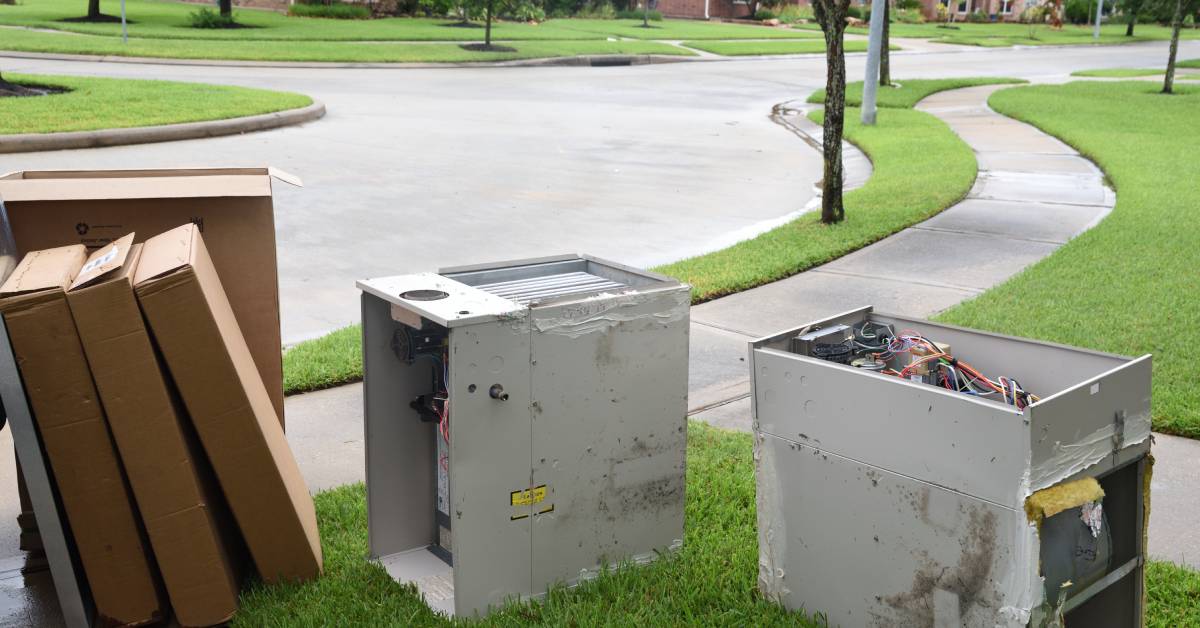 Two used gas furnace units and a stack of rectangular cardboard boxes sitting on the grass near the curb in a suburb.