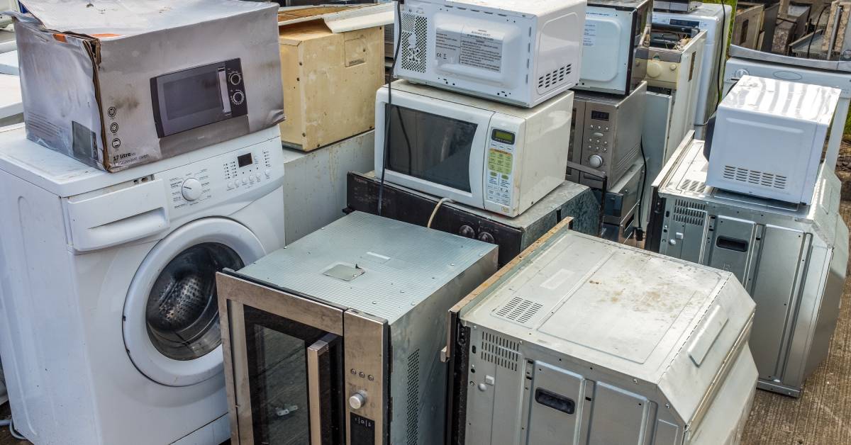 A large pile of broken residential appliances, including washing machines and microwaves, is sitting outside.