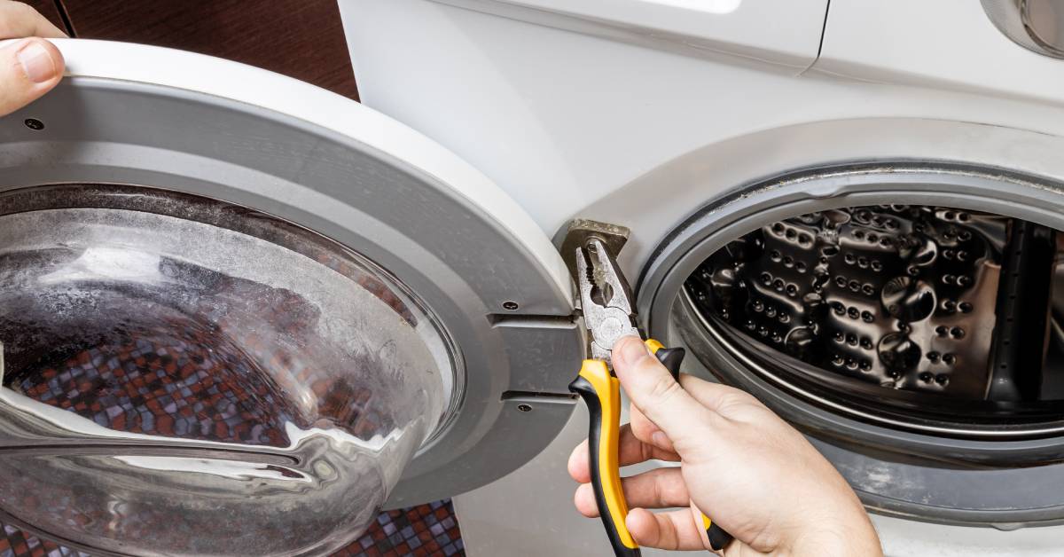 A hand holding black and yellow pliers, adjusting a screw on a washing machine door hinge while the door is open.
