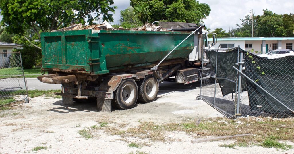 A truck carries a green roll-off dumpster from a construction site, filled with construction debris.