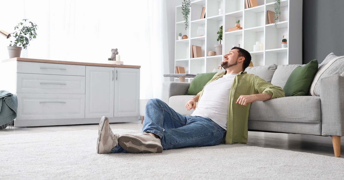 A man sitting on his living room floor, leaning against a gray couch with green pillows in front of white cubby shelves.