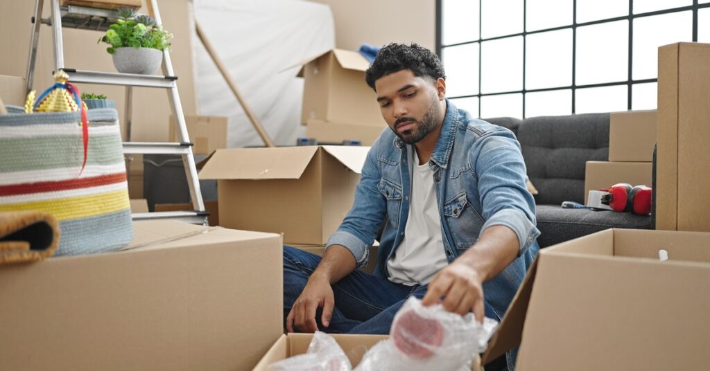 A person wearing a denim jacket pulls out a plastic wrap from one of the multiple cardboard boxes surrounding him.