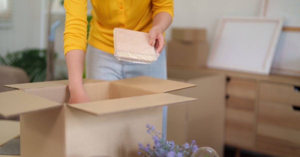 A person wearing a yellow shirt and gray pants is putting items into a cardboard box next to a lavender plant.