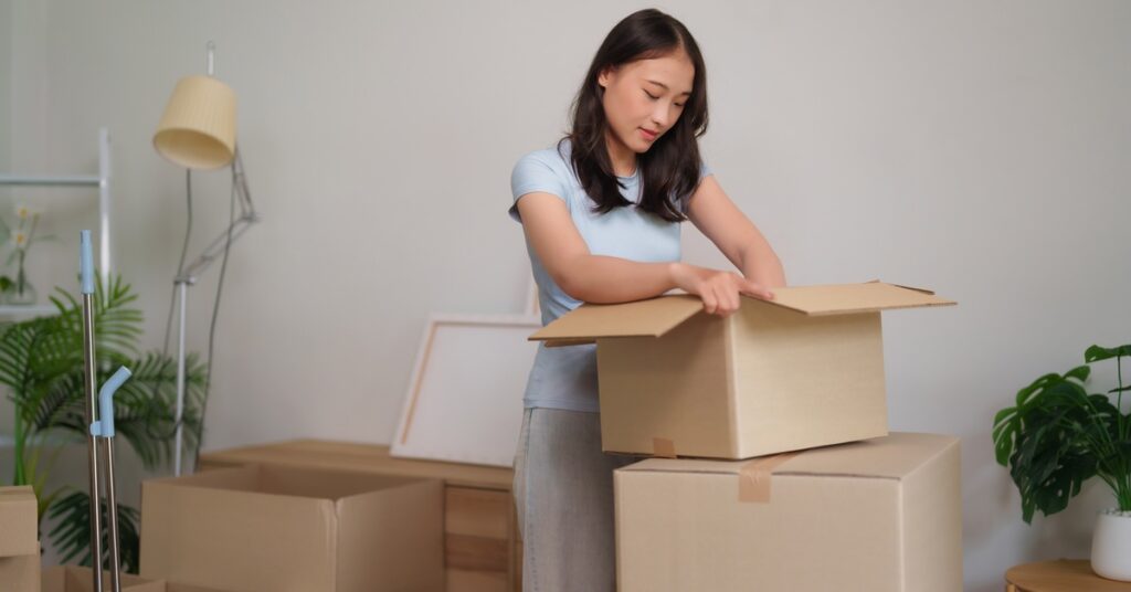 A woman wearing a light blue shirt looks through a smaller cardboard box sitting on top of a bigger cardboard box.