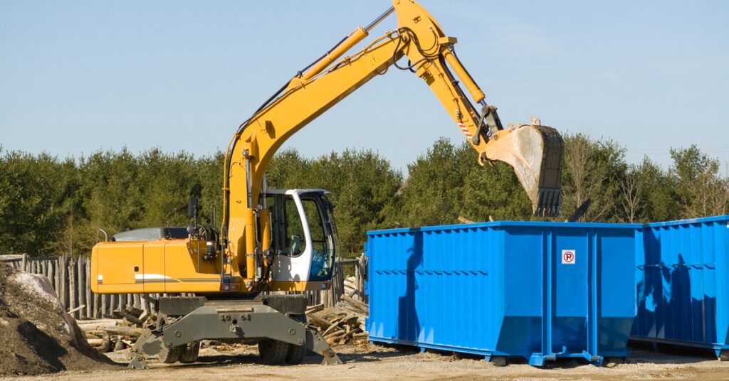 A heavy-duty demolition bulldozer's blade hovers above a blue dumpster with a no parking sign attached to it.