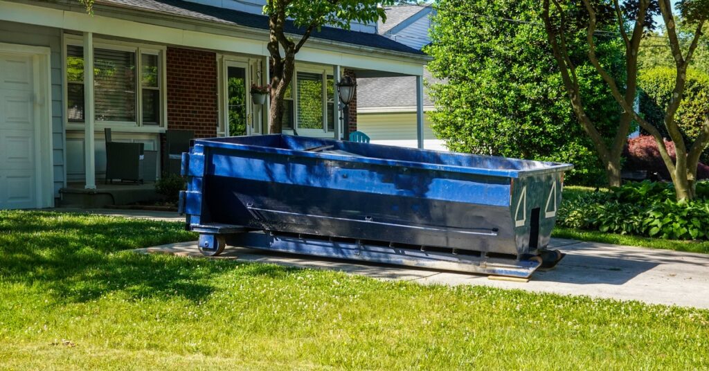 A large blue dumpster sits in the front yard of a red, residential house, surrounded by grass and trees.