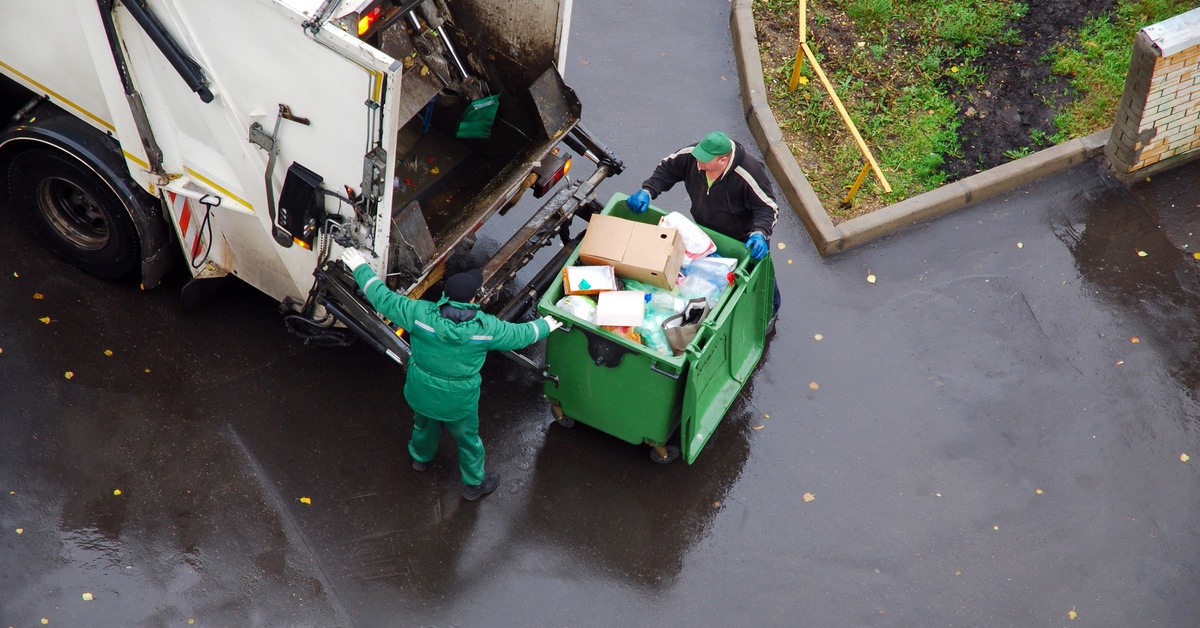 Two people push a front overload dumpster containing household rubbish across wet concrete into a garbage truck.