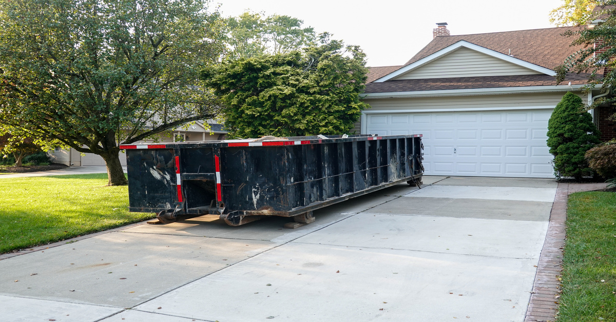 A black and red dumpster is parked on a concrete driveway, in front of a brown house and next to a tree.