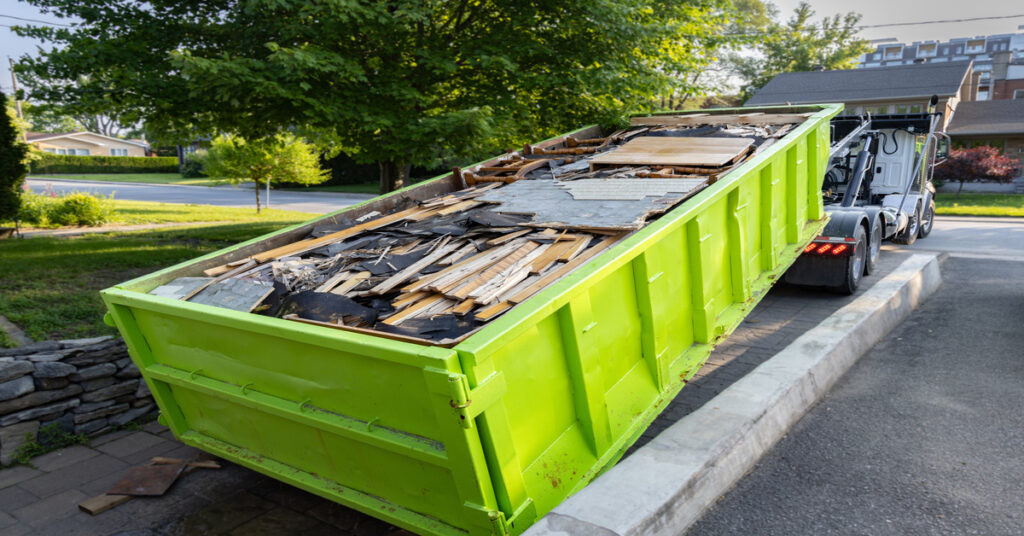 A lime green dumpster filled with construction materials is being loaded by a truck off a concrete driveway.