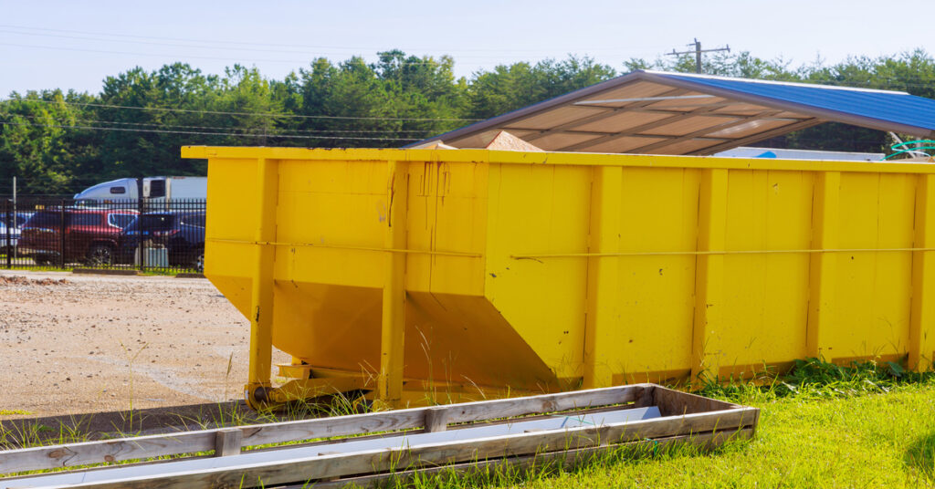 A bright, yellow dumpster filled with waste is parked on the grass, next to wooden planks for construction.