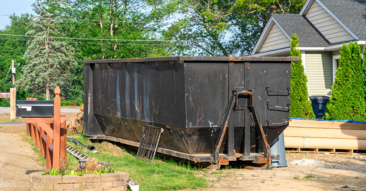 A black dumpster parked in front of a white house with a black and orange mailbox. Trees are in the background.