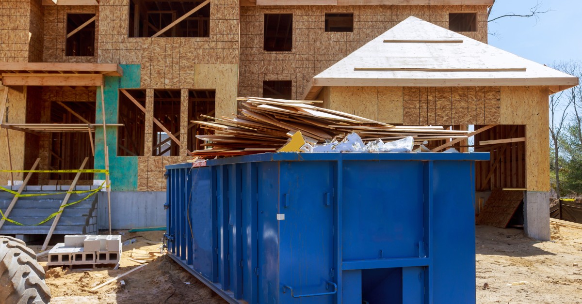 A blue dumpster is filled with building material waste and parked in front of house under construction.