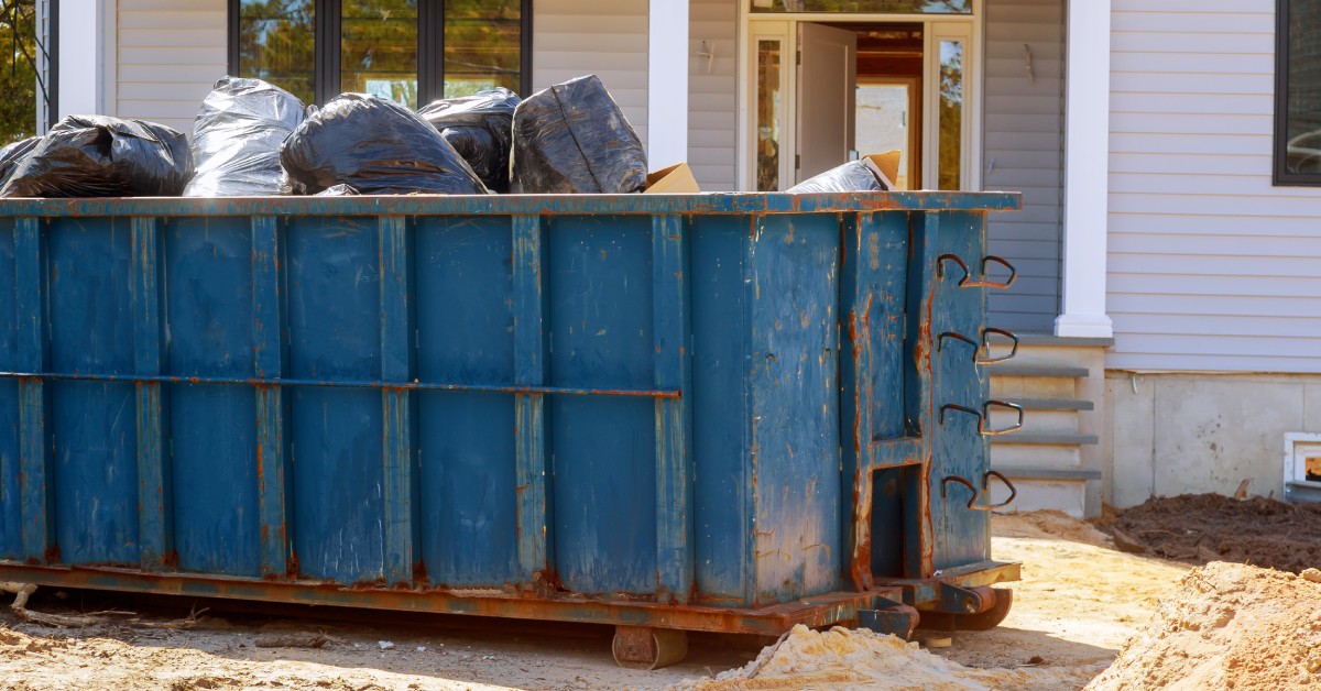 A blue dumpster filled with black garbage bags is parked in front of a gray house that is under construction.