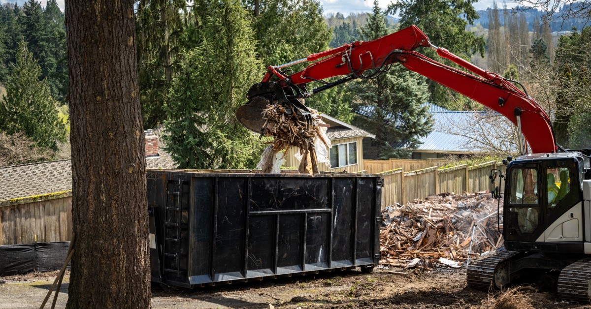 A red excavator uses a jawbone bucket to transfer construction waste into a black iron dumpster, next to a tree.