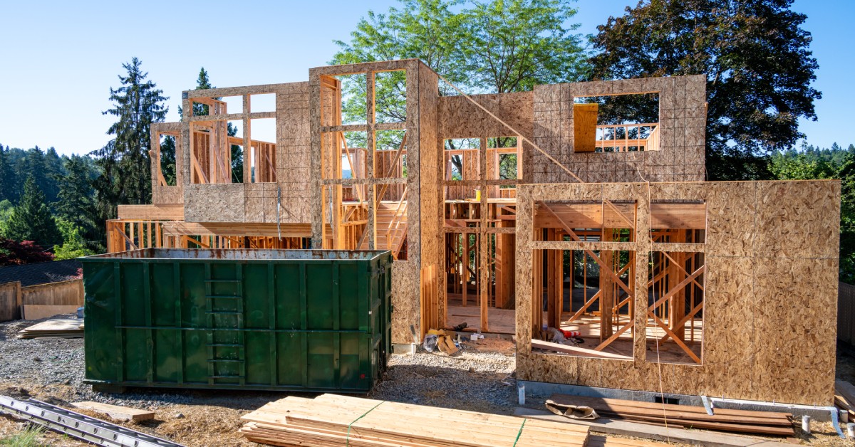A dark green dumpster sits in front of a two-story house under construction, surrounded by wooden planks and materials.