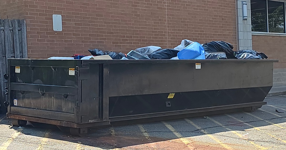 A large black roll-off dumpster filled with garbage bags and trash is parked next to a red brick building.
