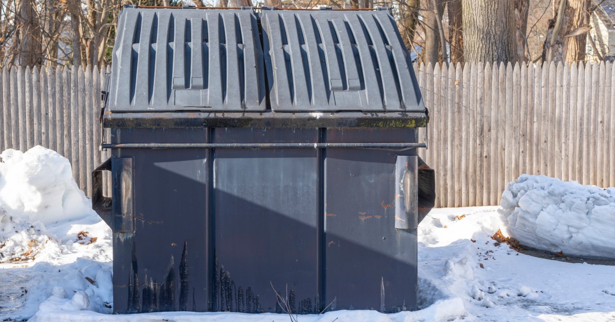 A commercial black-and-gray dumpster in a parking lot, surrounded by snow, with wooden fences and trees behind it.