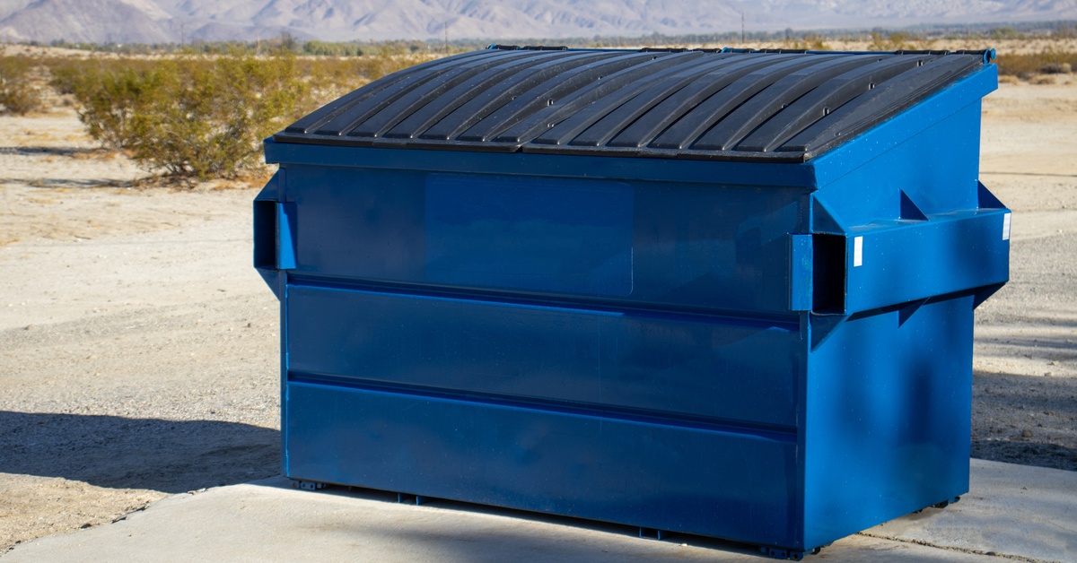 A large blue commercial dumpster sits outdoors on concrete. Behind it are mountainous ranges and a desert.