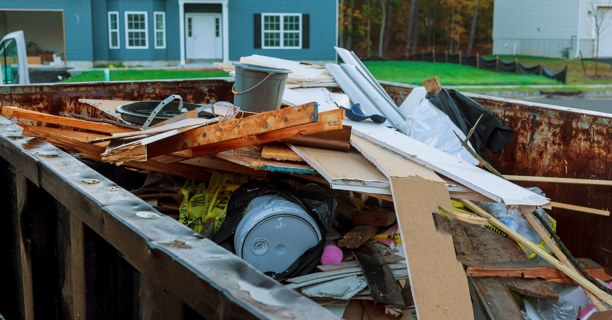A black, roll-off dumpster filled with different kinds of waste and rubble. Behind it is a blue, multi-story house.
