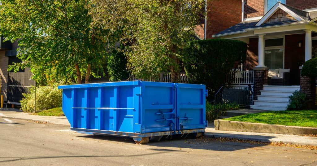 A small, blue roll-off dumpster sits on the sidewalk in front of a red brick house, next to greenery.