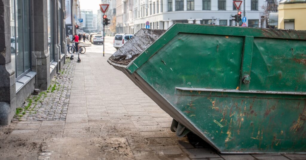 A green, dumpster filled with soil and debris sit on an urban sidewalk in the city, next to commercial buildings.